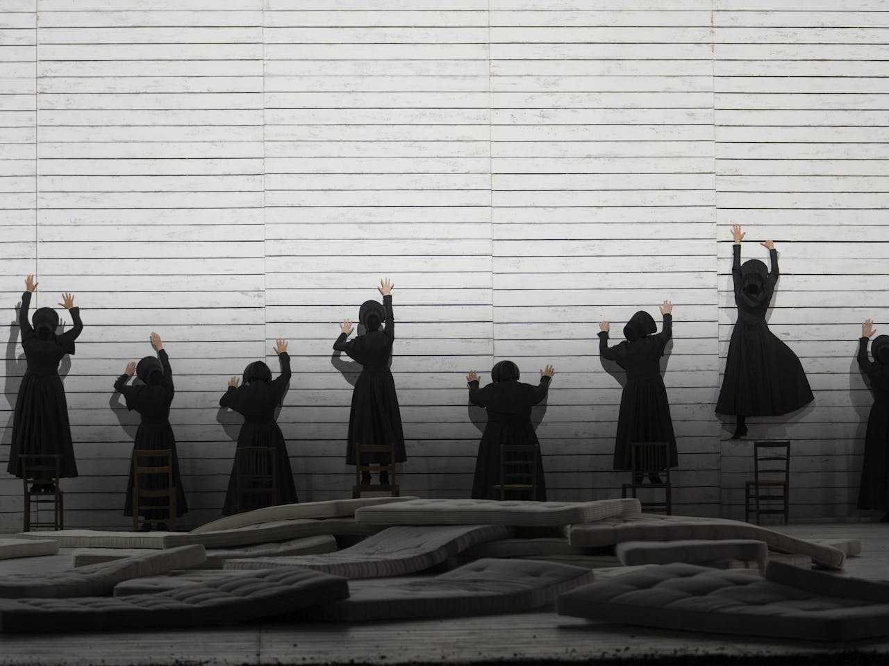 A group of women dressed in long black dresses attempt to climb up a sheer white wall. They have their backs to the camera.