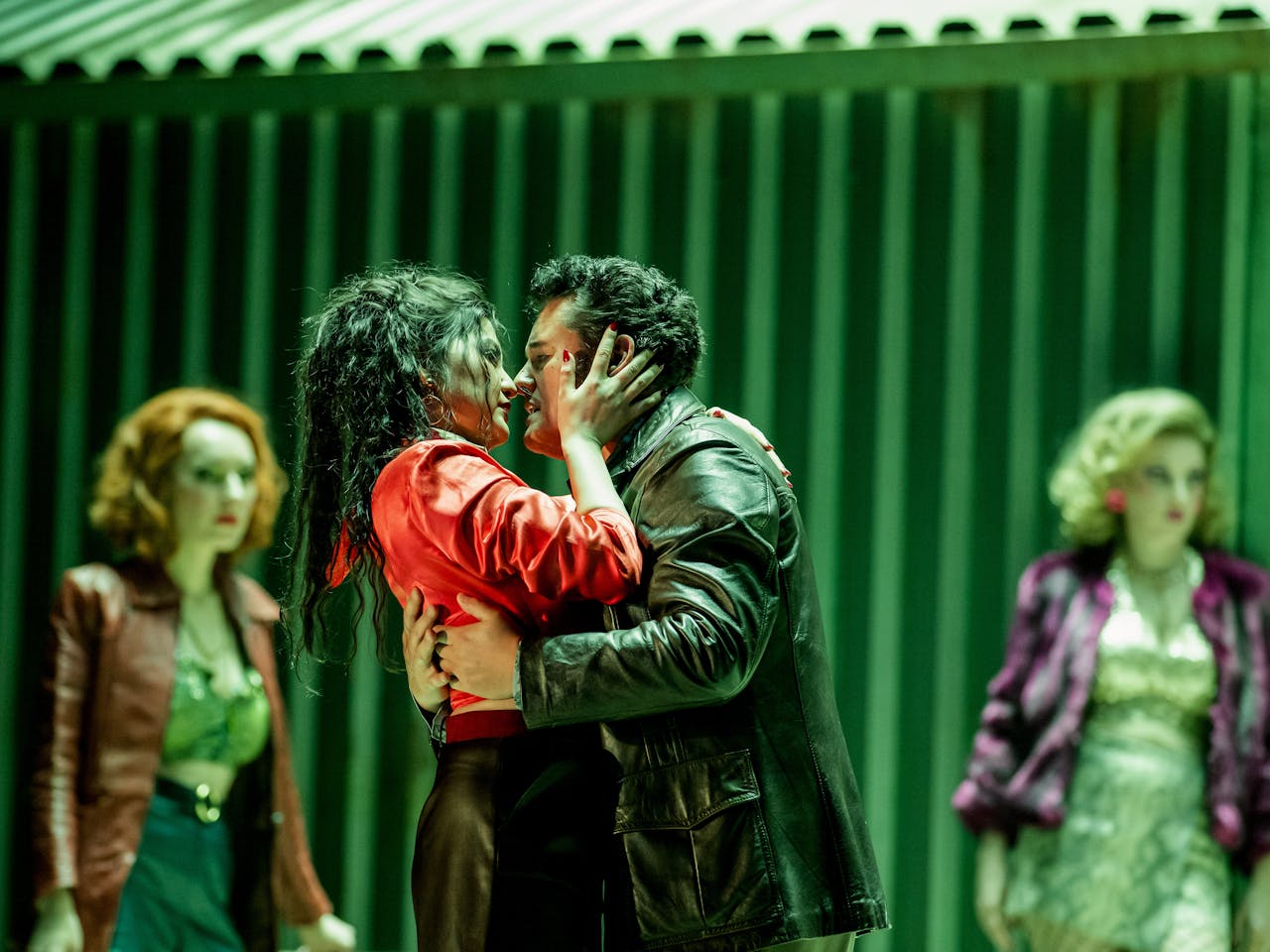 An opera singer with long black hair wearing red holds the head of another opera singer wearing a leather jacket in a passionate embrace. Two performers stand behind them looking on. 