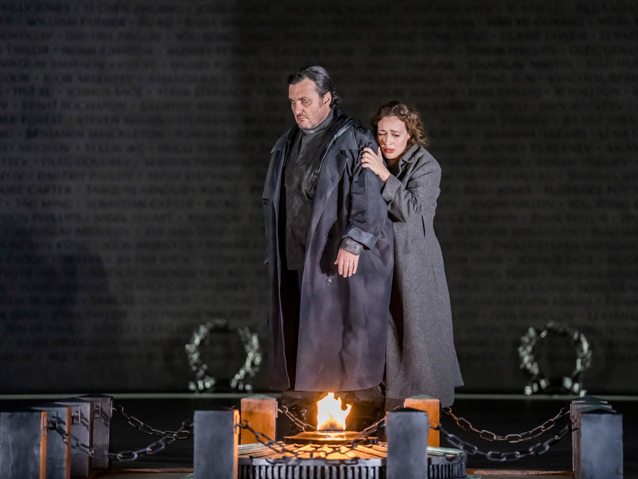 Two characters embrace, one behind the other as they face an open flamed fire. They are both wearing long, heavy grey overcoats