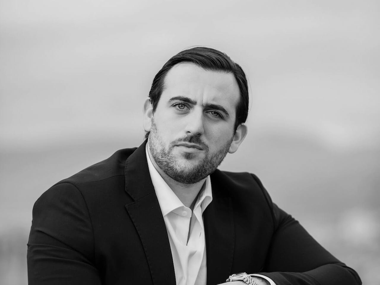 A black and white headshot of a person wearing a black jacket and white shirt. 