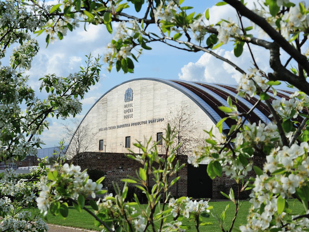 A building with a rounded roof, a brick wall and short-cut grass sits in the foreground and is framed with flowing flowers. 