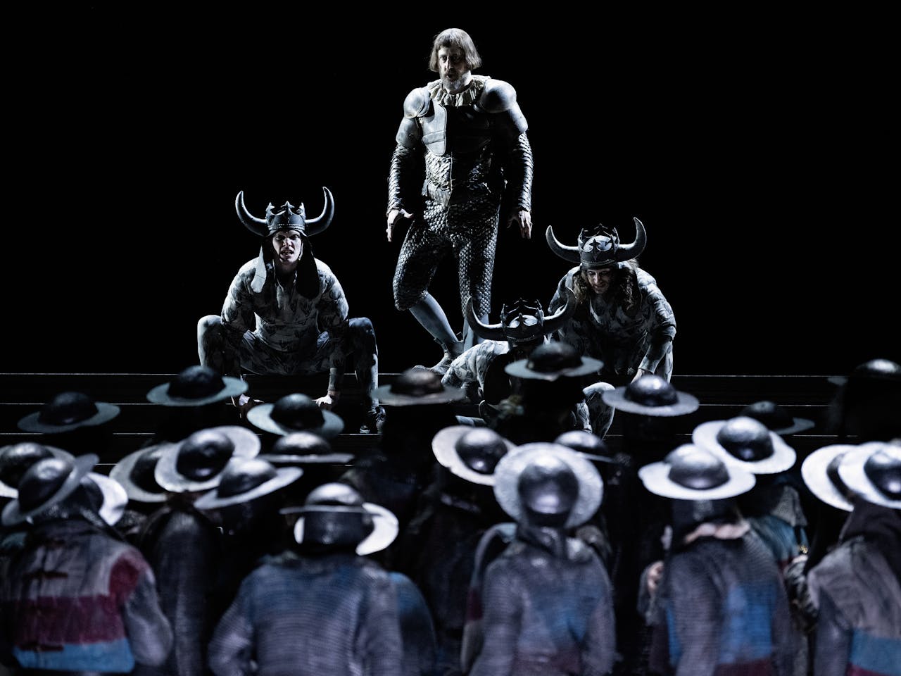 The Royal Opera perform Il Trovatore. A man stands against a black background in armour, looking out over a troop of soldiers in metal helmets.