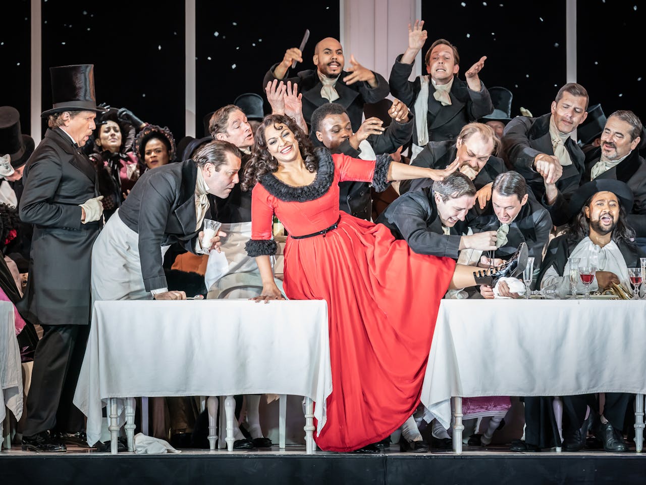 A woman in a red dress with black fur lining sits on a white draped table with several men around. 