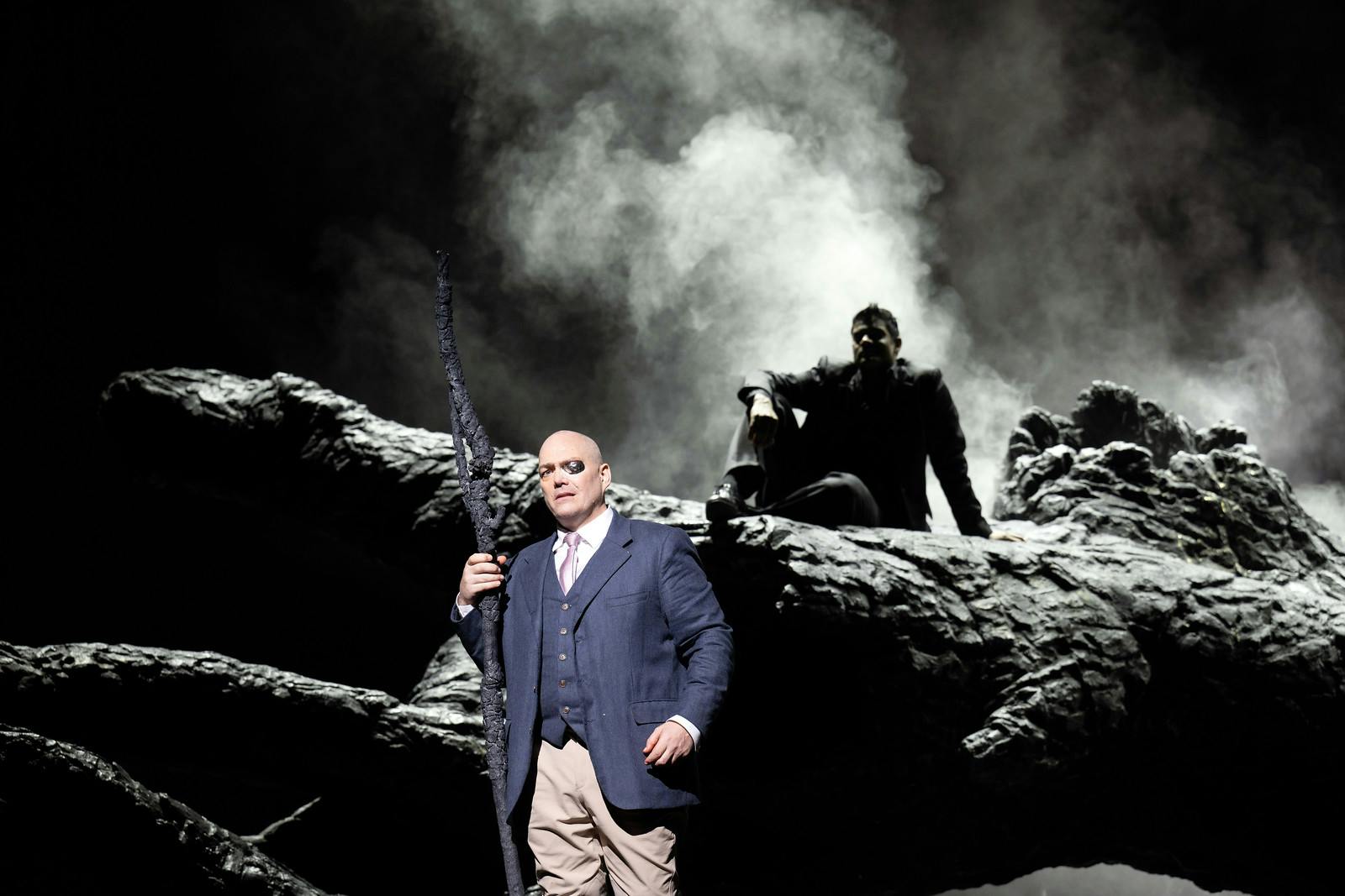 A man dressed in a blue suit wearing an eyepatch performs onstage in The Royal Opera production, Das Rheingold.