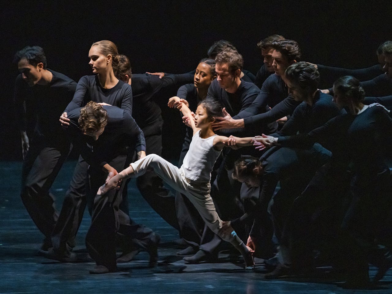 Several dancers wearing black hold onto a small child dressed in white, who appears to be running