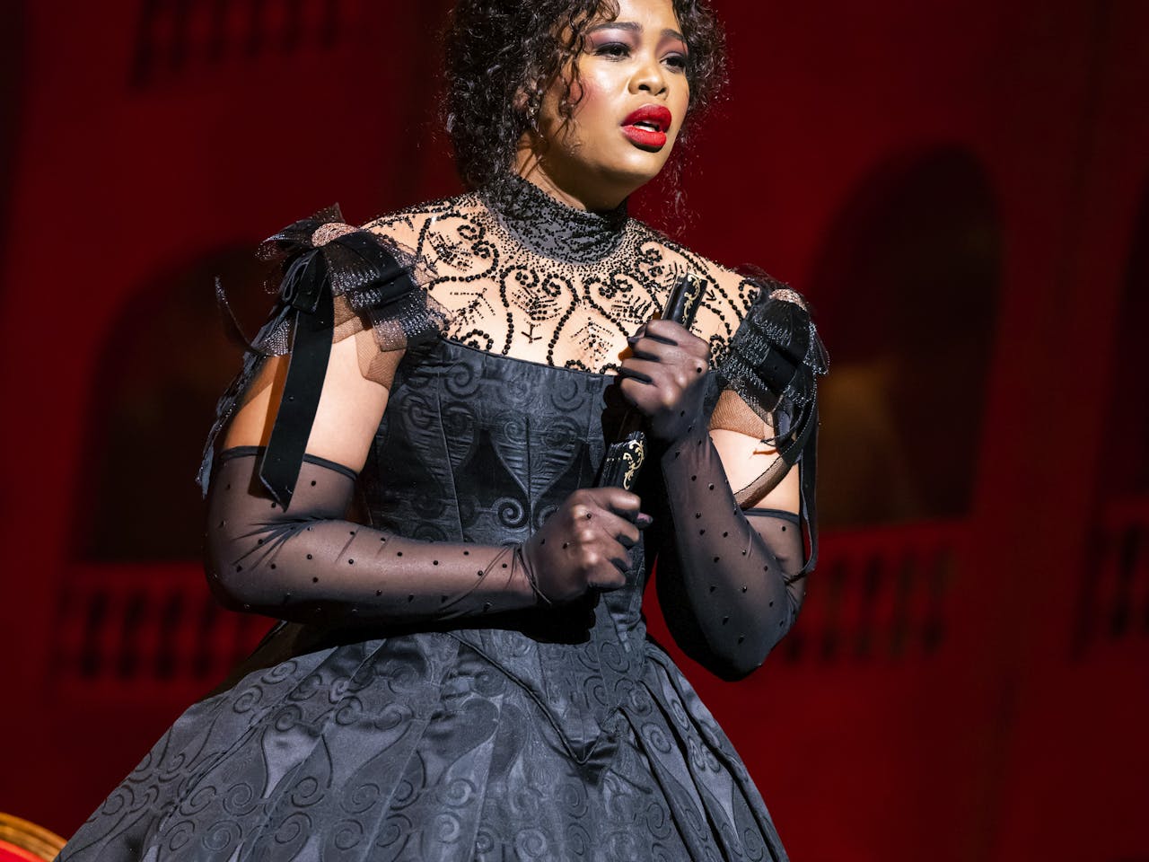 A woman in a black dress holding a fan looks out onto the audience from the stage as she sings.
