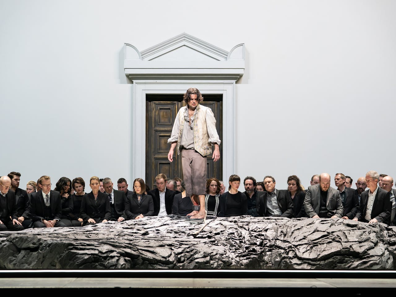 A performer stands on a raised rock with their ankle shackled. Behind them a group of people look on with serious expressions on their face