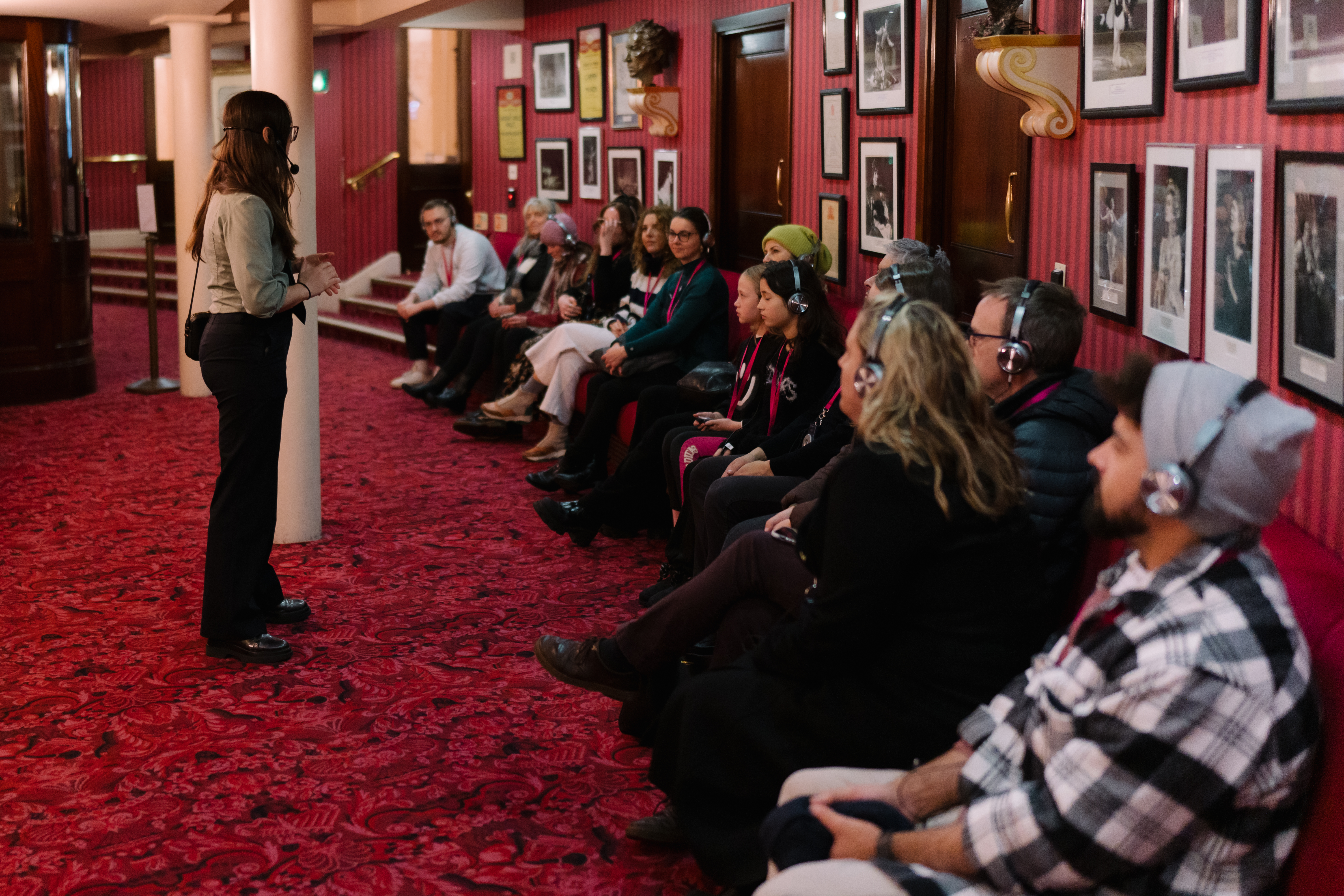 A guide speaks to a seated tour group wearing headsets in a curved room with red carpet and framed photos on the walls.