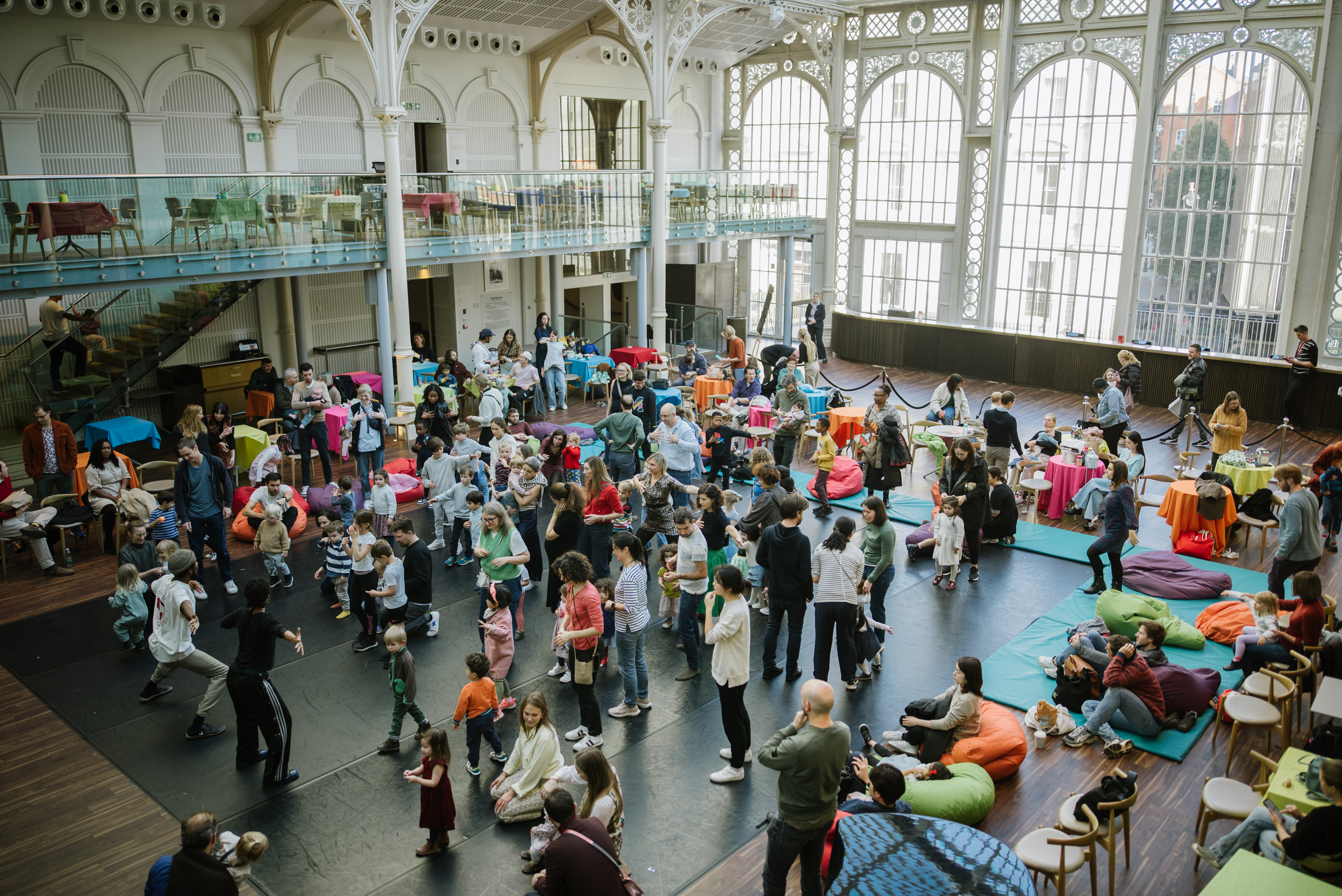 A large group of people gathered in a brightly lit room with vaulted ceilings and white ironwork windows, they are engaged in conversation and activities.