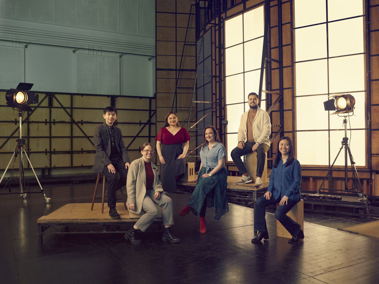 A group of performers stand and sit in the middle of the backstage are. The backstage area has stage lights in metal frames and a raised wooden platform