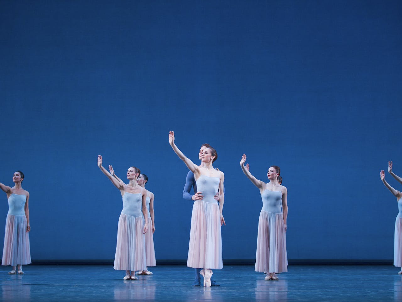 10 ballet dancers stand on a blue-set stage holding their right arms in the air. In the centre, there are 5 dancers and either side of hte stage are three dancers - all are wearing skirts. The dancer at the front is being held by the waist by another dancer wearing blue trousers and top.

