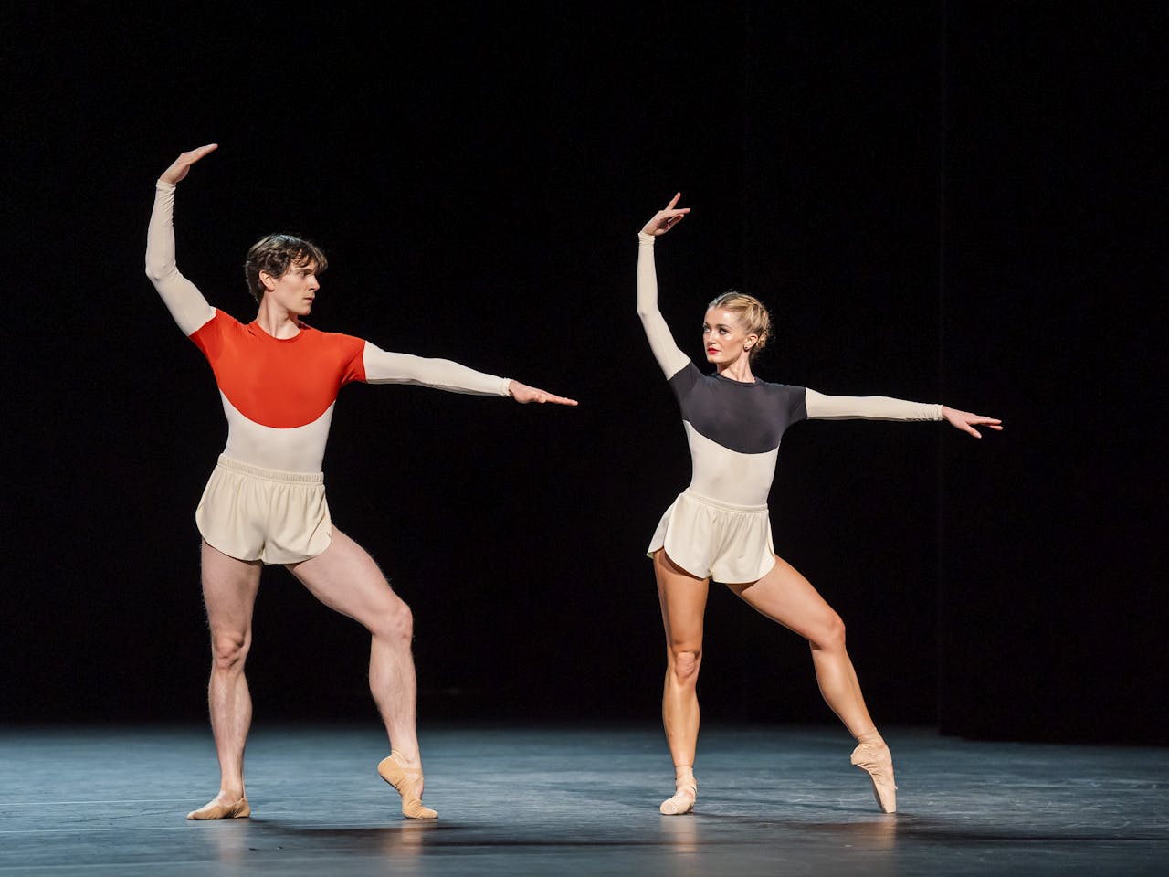 Two ballet dancers wearing shorts and a leotard stand on stage facing the audience. Their left arms are stretched straight and their right arm is curved above their heads. They are principal dancers of The Royal Ballet William Bracewell and Anna Rose O'Sullivan.
