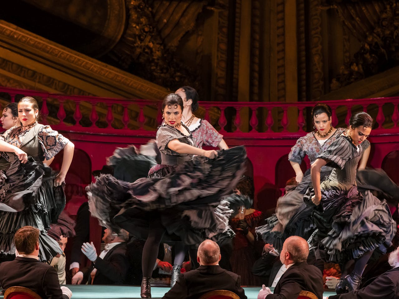 Three flamenco dancers in black dresses perform on stage with 3 male flamenco dancers behind them.