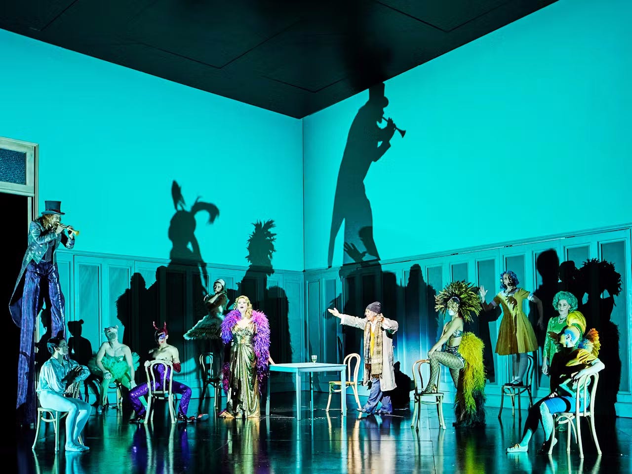 The stage is filled with a variety of characters in bright costumes and outfits - a singer wearing a gold dress and purple feather boa stands in the centre while people on stilts playing trumpets are lit creating shadows.