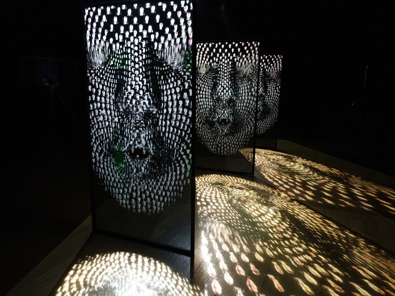Three black screens have images of pixilated faces projected onto them and shadows are formed on the floor around.