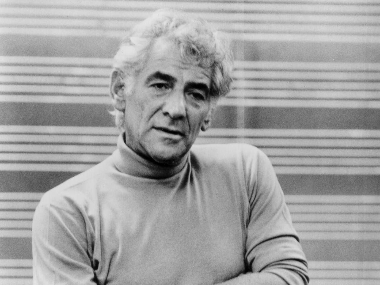 Leonard Bernstein stands facing the camera with a neutral expression. He is wearing a light turtle neck and jeans. His arms are folded.