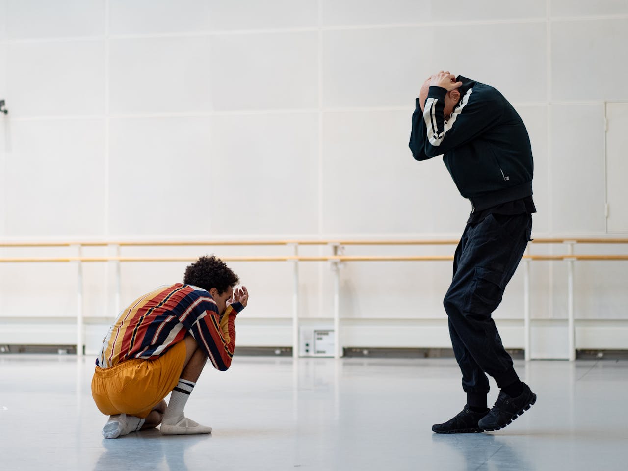 Two dancers rehearse in a white ballet studio. One is dressed in orange and crouches on the floor, the other is wearing a black tracksuit. Both have their heads in their hands. 