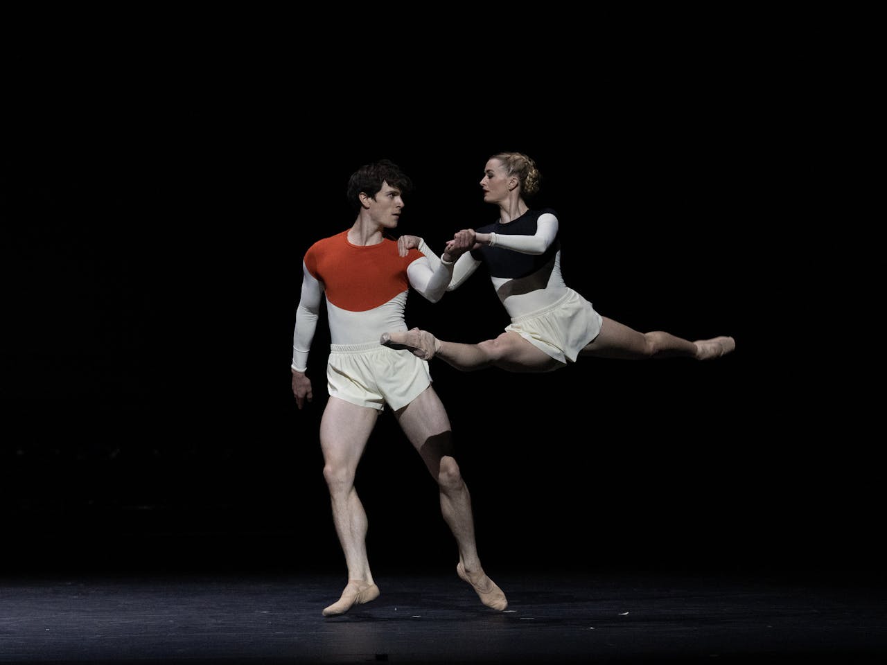 Two dancers, William Bracewell and Anna Rose O'Sullivan, perform on a dark stage. Anna Rose O'Sullivan is in mid-air holding a splits. William Bracewell holds her hand. They are wearing white shorts and white, red and blue long sleeve shirts.