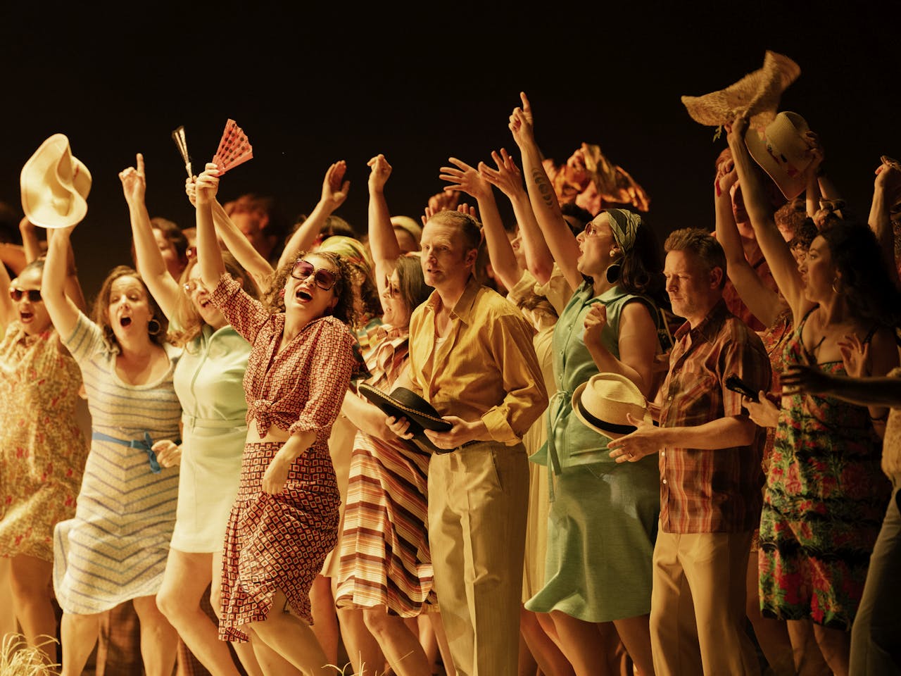 A group of performers who make up the Chorus of The Royal Opera House dance on stage with their arms raised high. They are all wearing patterned and colourful clothing.