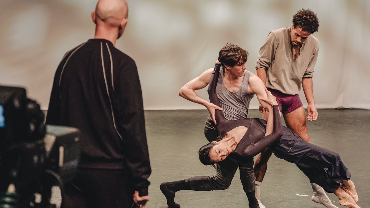 A film camera in the corner overlooks the back of Resident Choreographer Sir Wayne Mcgregor as he watches three dancers of The Royal Ballet rehearse in the centre of a studio. One dancer is leaning against another and the third is performing behind them. 